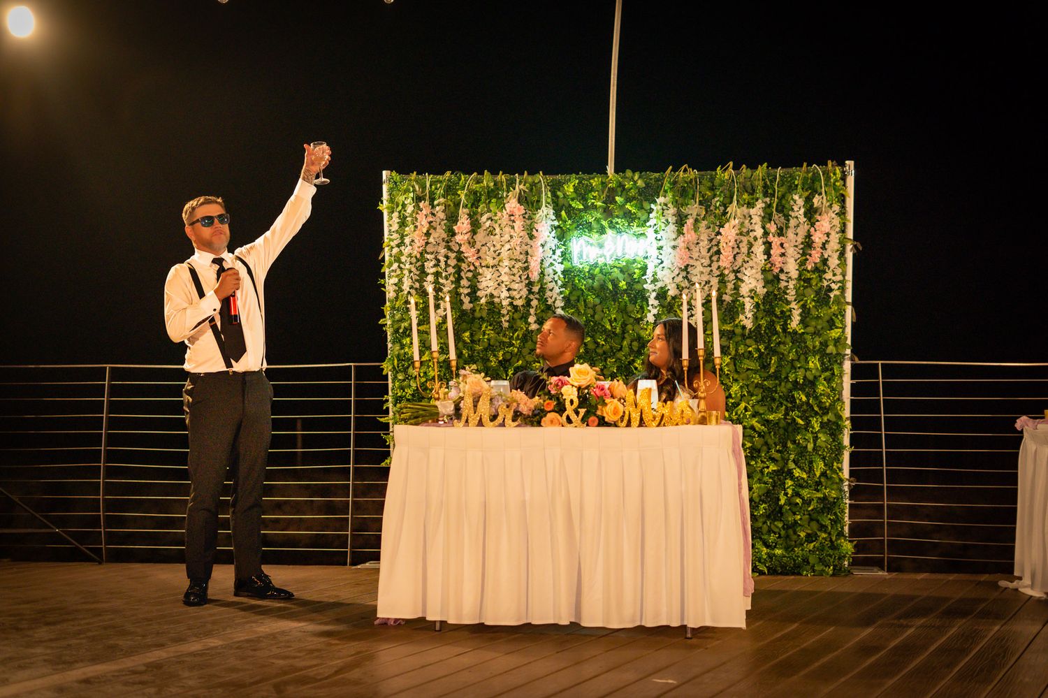 A man in sunglasses raises his hand at an outdoor evening event near a decorated table with greenery backdrop.
