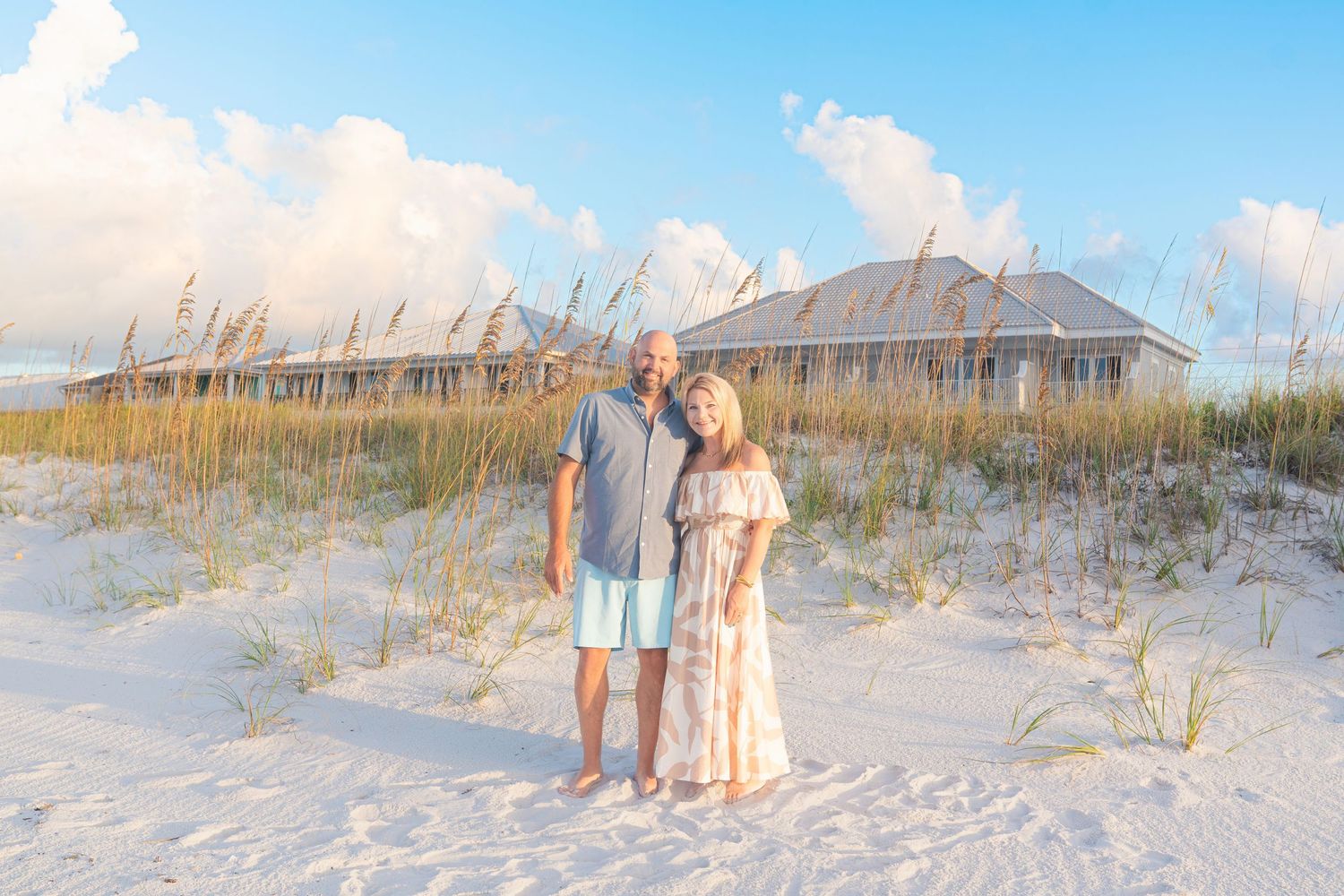 Couple smiling together on white sand beach with sea oats and coastal cottage in background at golden hour.