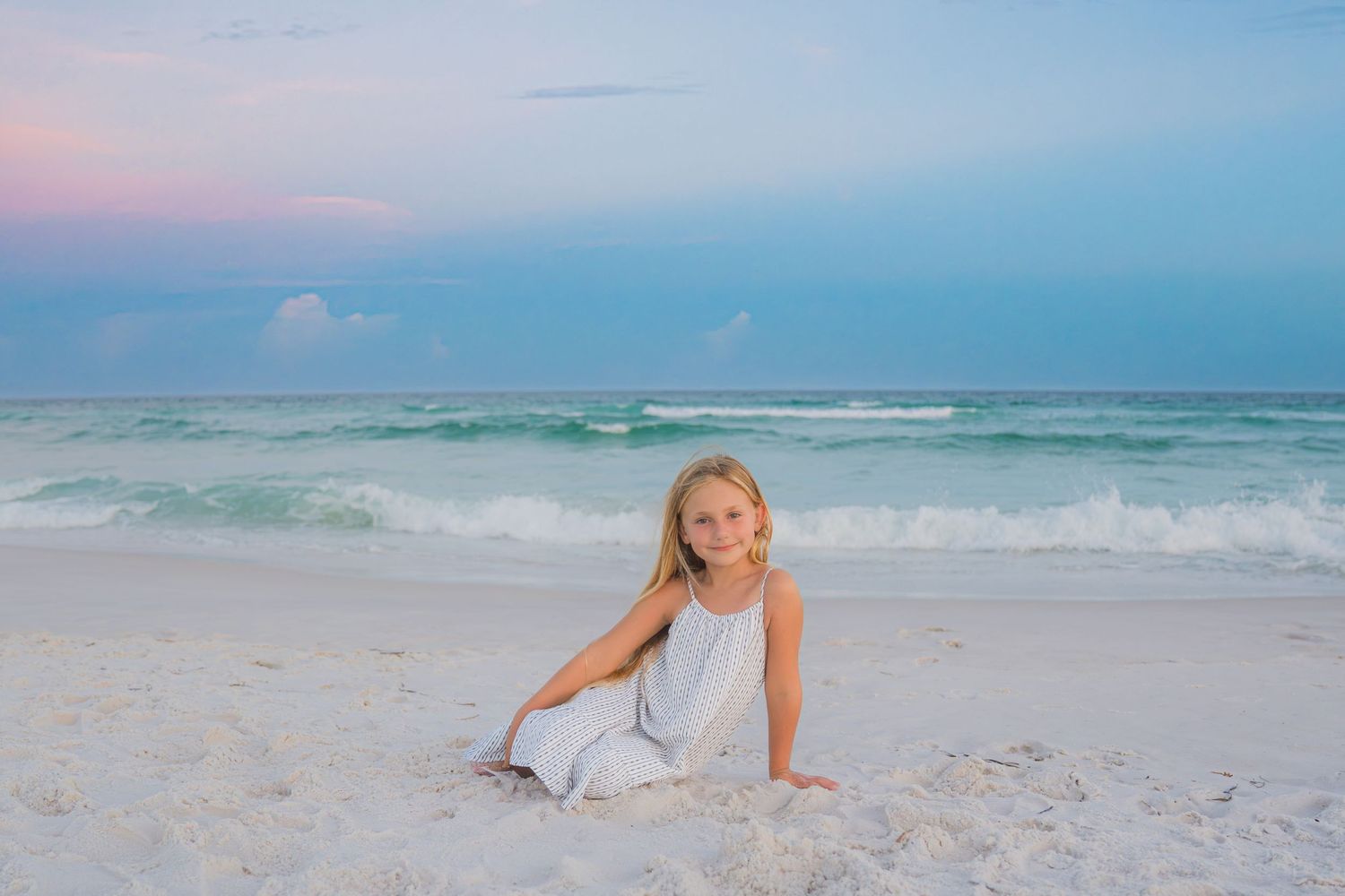 Young girl in white dress sitting on white sandy beach at sunset with turquoise ocean waves behind her.