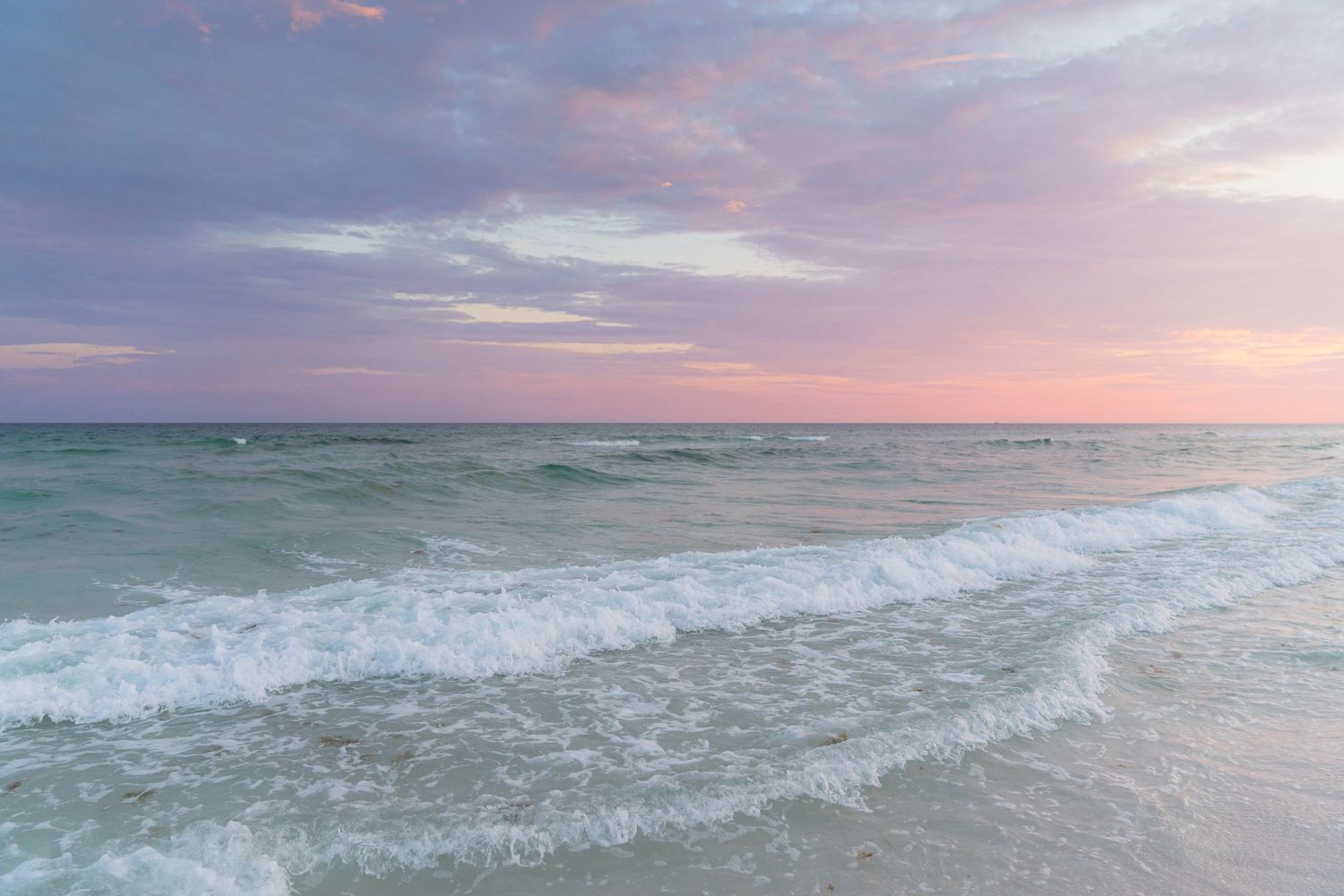 Pastel pink and purple sunset sky over calm turquoise ocean waves gently rolling onto a sandy beach.