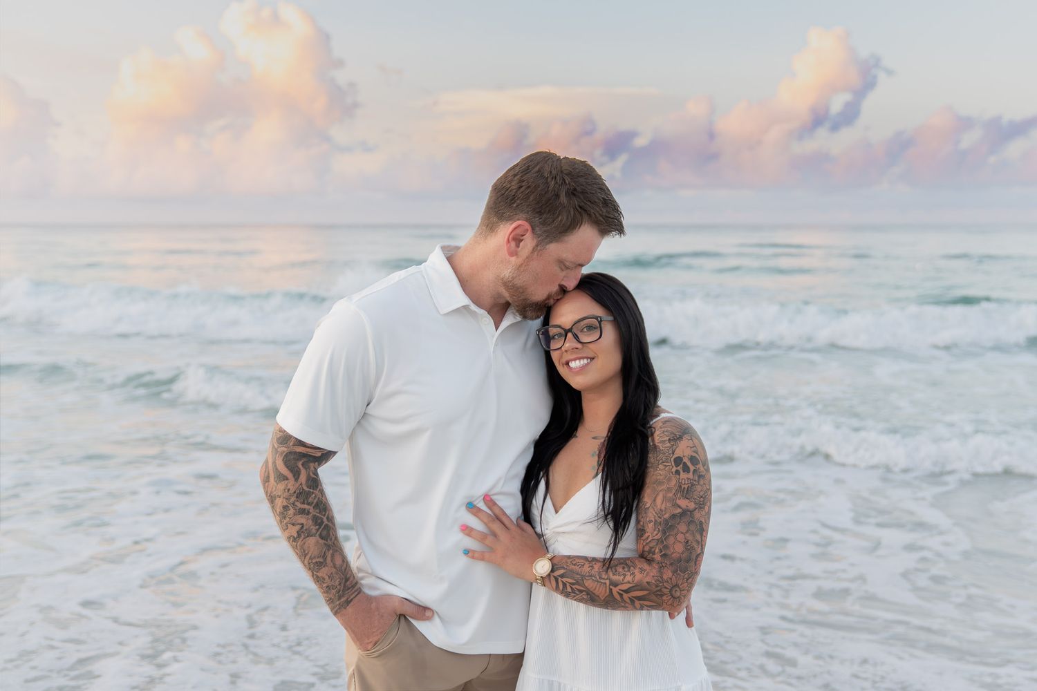 Tattooed couple sharing a romantic moment on a beach with soft pastel sky and ocean waves in background.