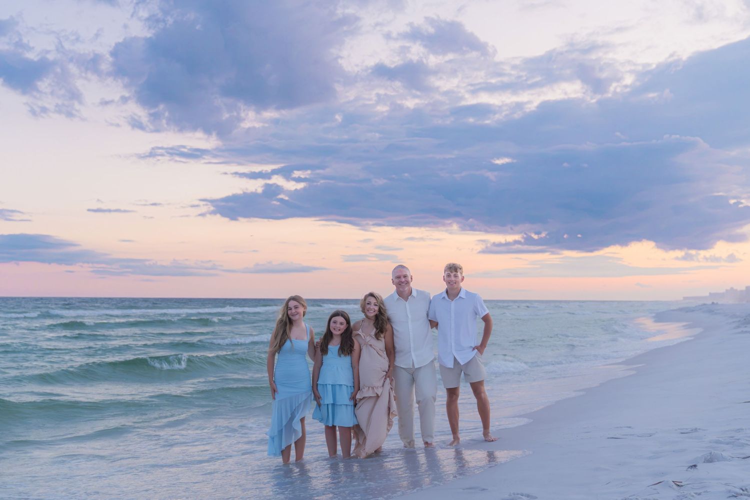 Family of five posing on a sandy beach at sunset with pastel skies and turquoise ocean waves behind them.