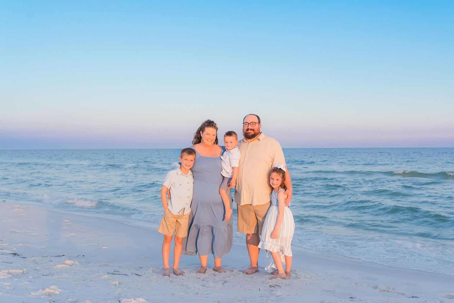 Family of five posing together on a sandy beach at sunset with calm ocean waves in the background.
