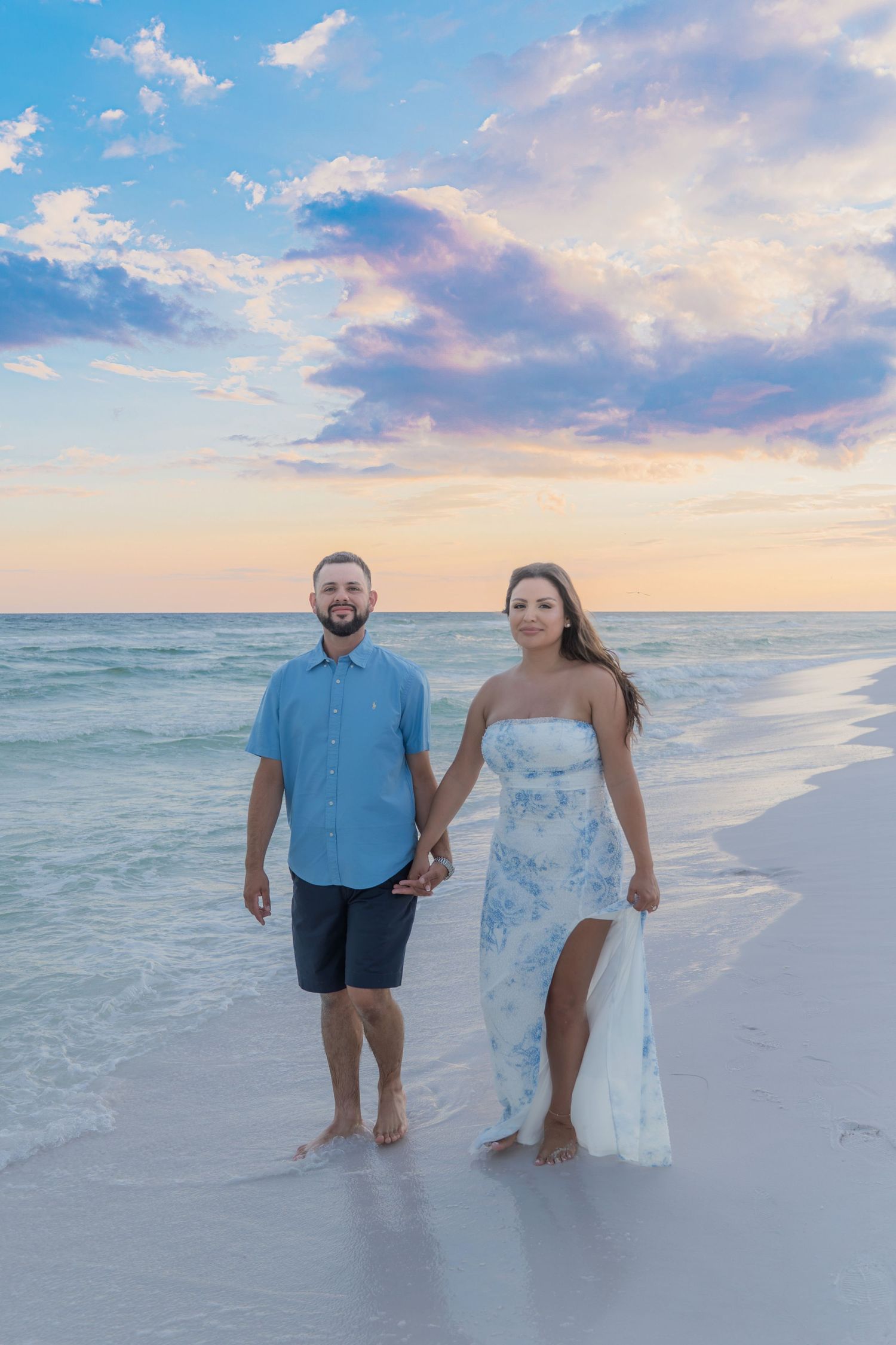 Smiling couple holds hands on white sand beach at colorful sunset, woman in floral dress and man in blue shirt.