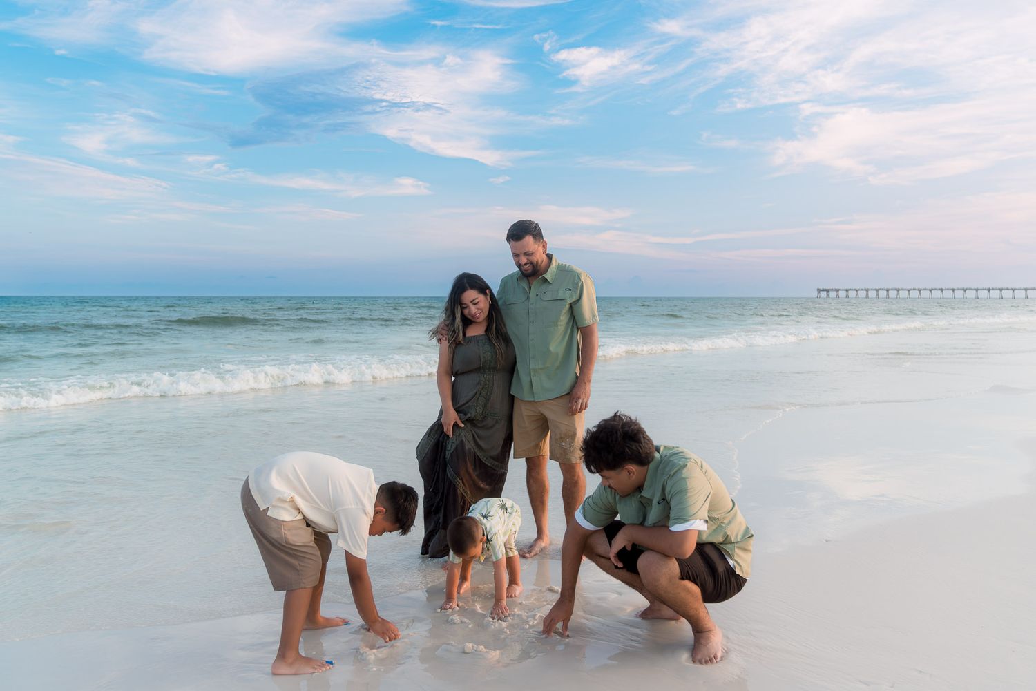 Family of five playing at the water's edge on a white sandy beach with a pier in the distance.