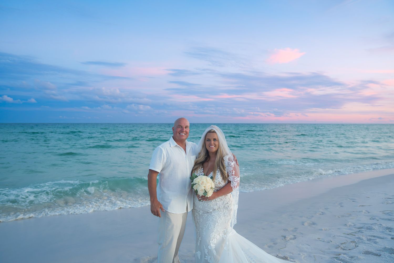 Bride and groom smiling together on a beach at sunset with turquoise water and pink sky.