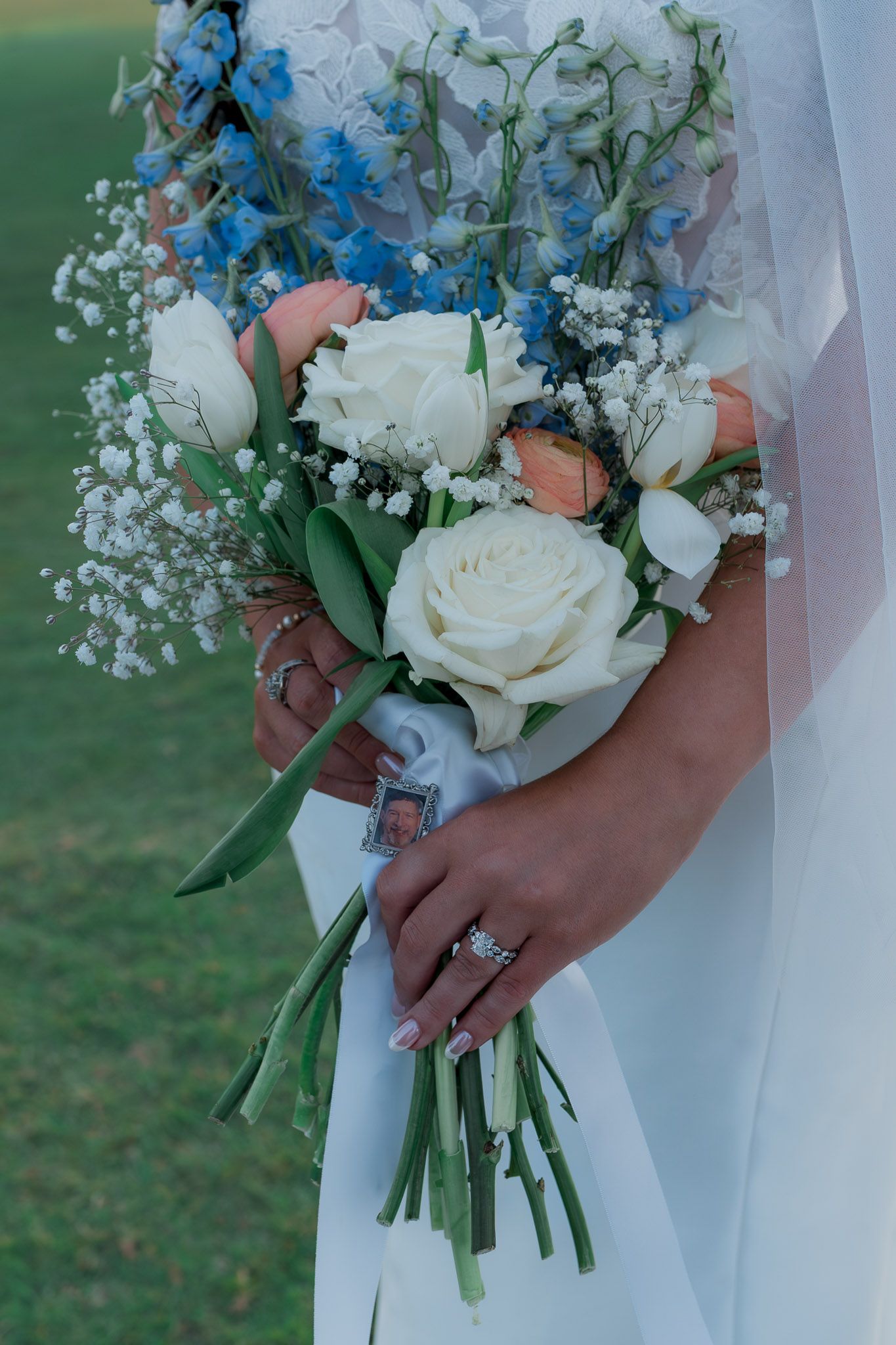 Bride holding elegant wedding bouquet with white roses, baby's breath, and blue flowers, wearing diamond rings.