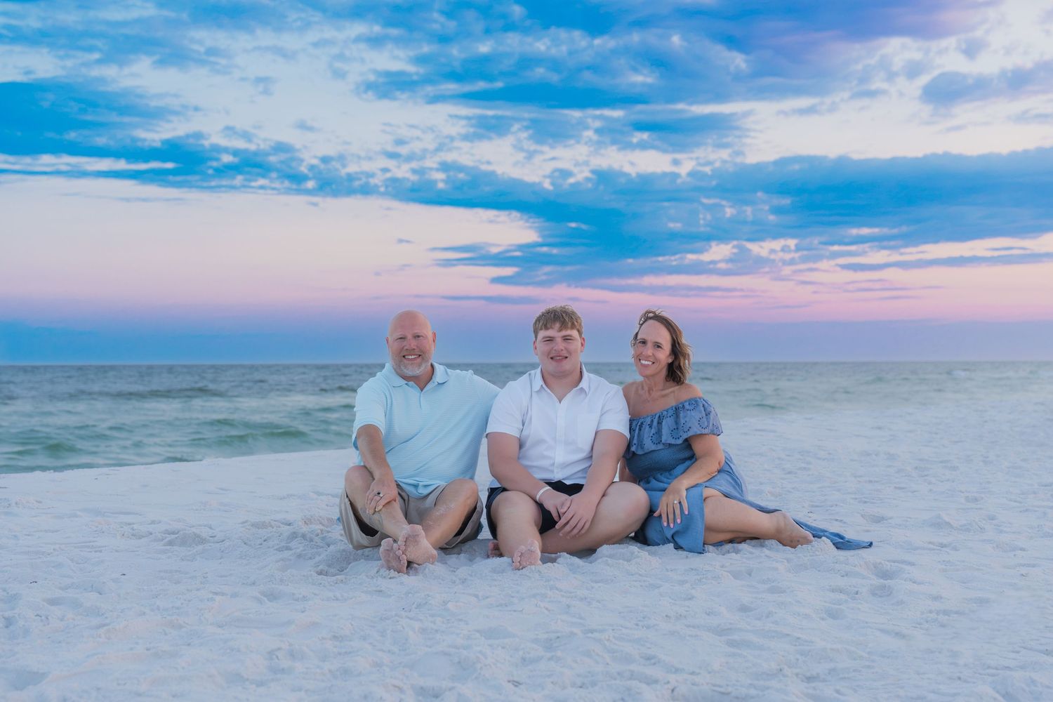 Family of three sitting on white sand beach at sunset with pink and blue sky backdrop.