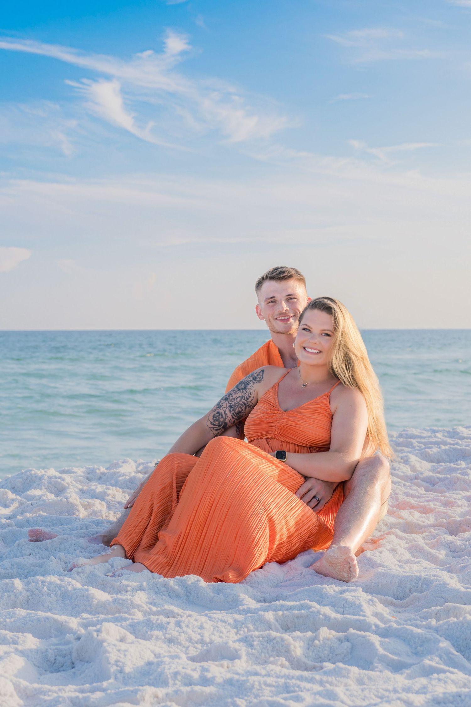 Couple in matching orange outfits sitting together on white sand beach with turquoise ocean and blue sky backdrop.