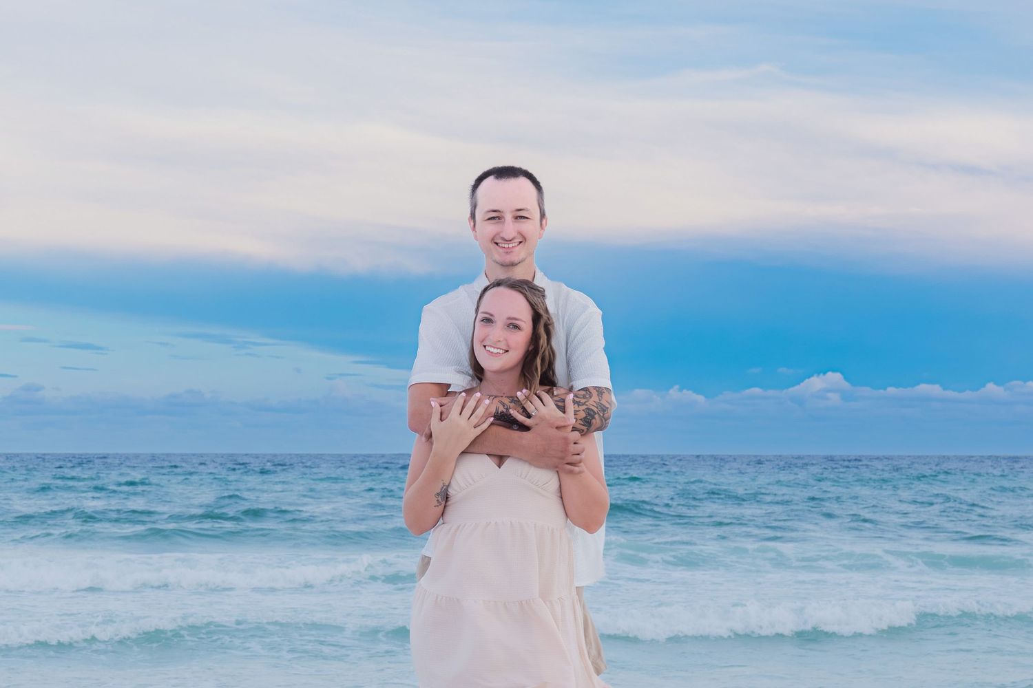 Couple embracing on a beach at sunset, woman in white dress, man with tattoos, turquoise ocean waves behind them.