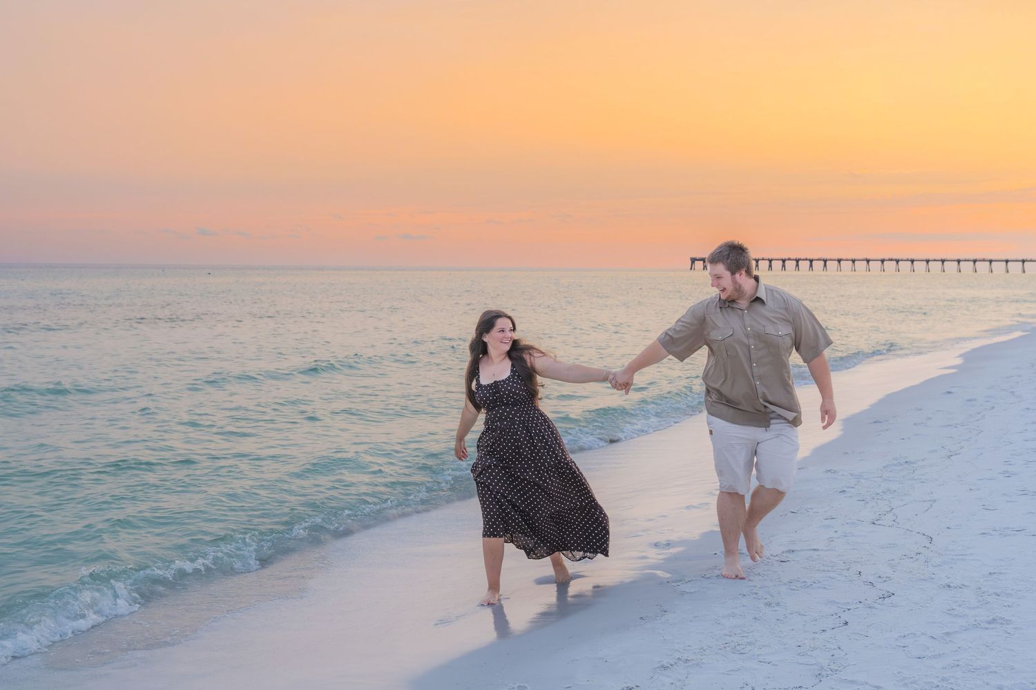 Couple holding hands walking on white sand beach at sunset with pier in background.