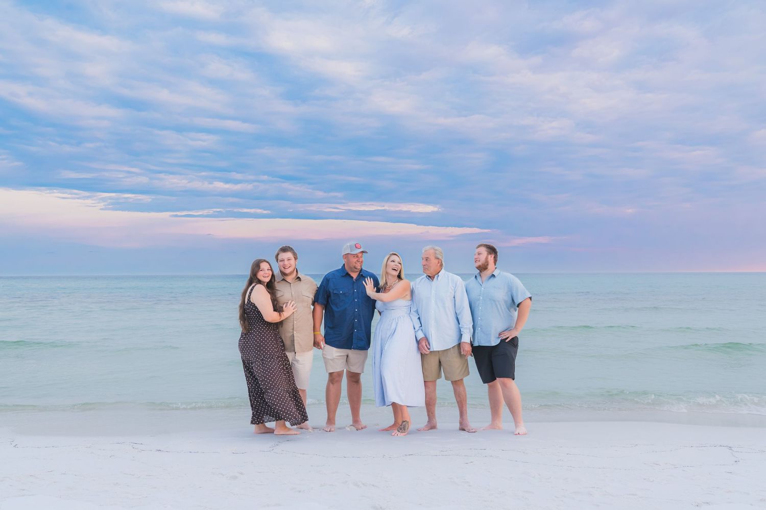Six people pose together on a white sand beach at sunset with turquoise water and pastel sky behind them.