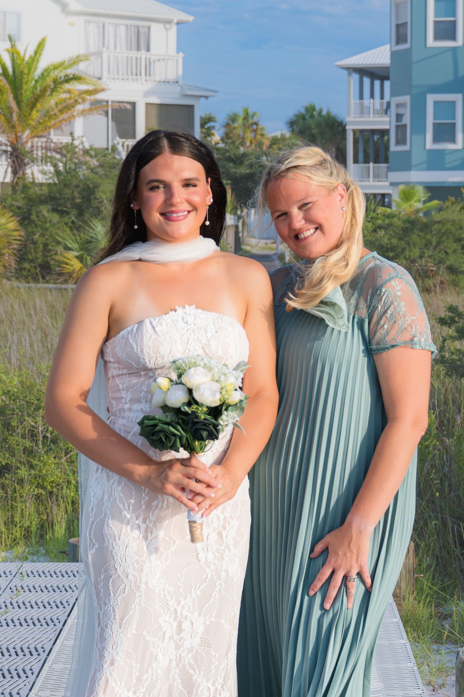 Bride in white lace gown holding bouquet poses with bridesmaid in sage green dress at beach wedding.