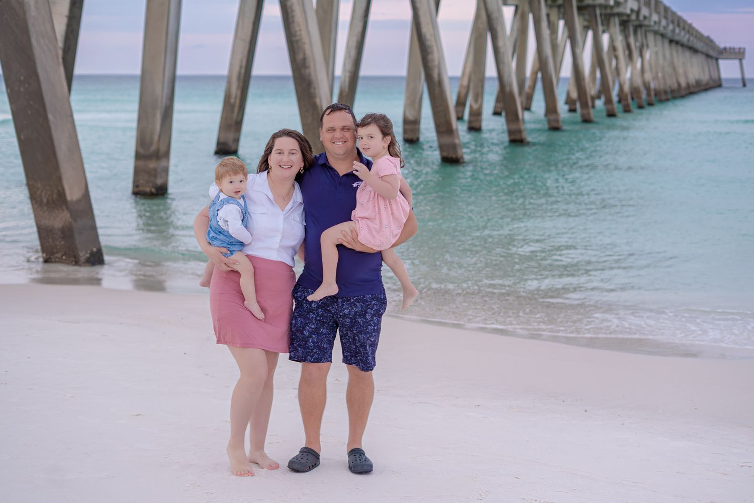 Family of four posing on a white sand beach near a wooden pier with turquoise water at sunset.
