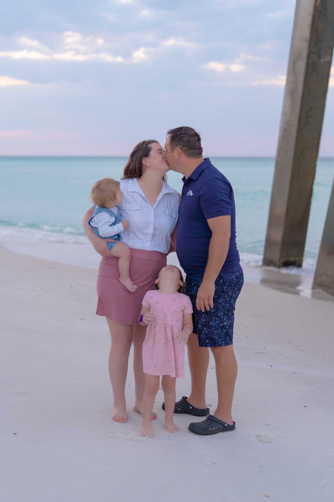 Family of four with two young children kissing on a white sand beach near a pier at sunset.