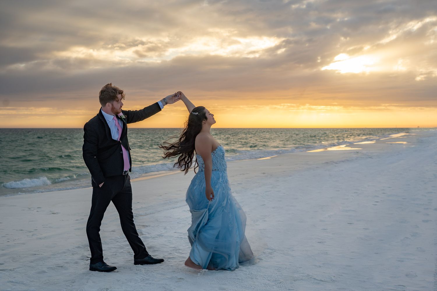 Couple dancing on a white sand beach at sunset, man in black tuxedo twirling woman in flowing blue gown.