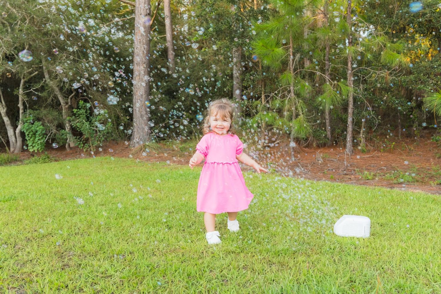 Young girl in pink dress playing with bubbles in a green backyard surrounded by trees.