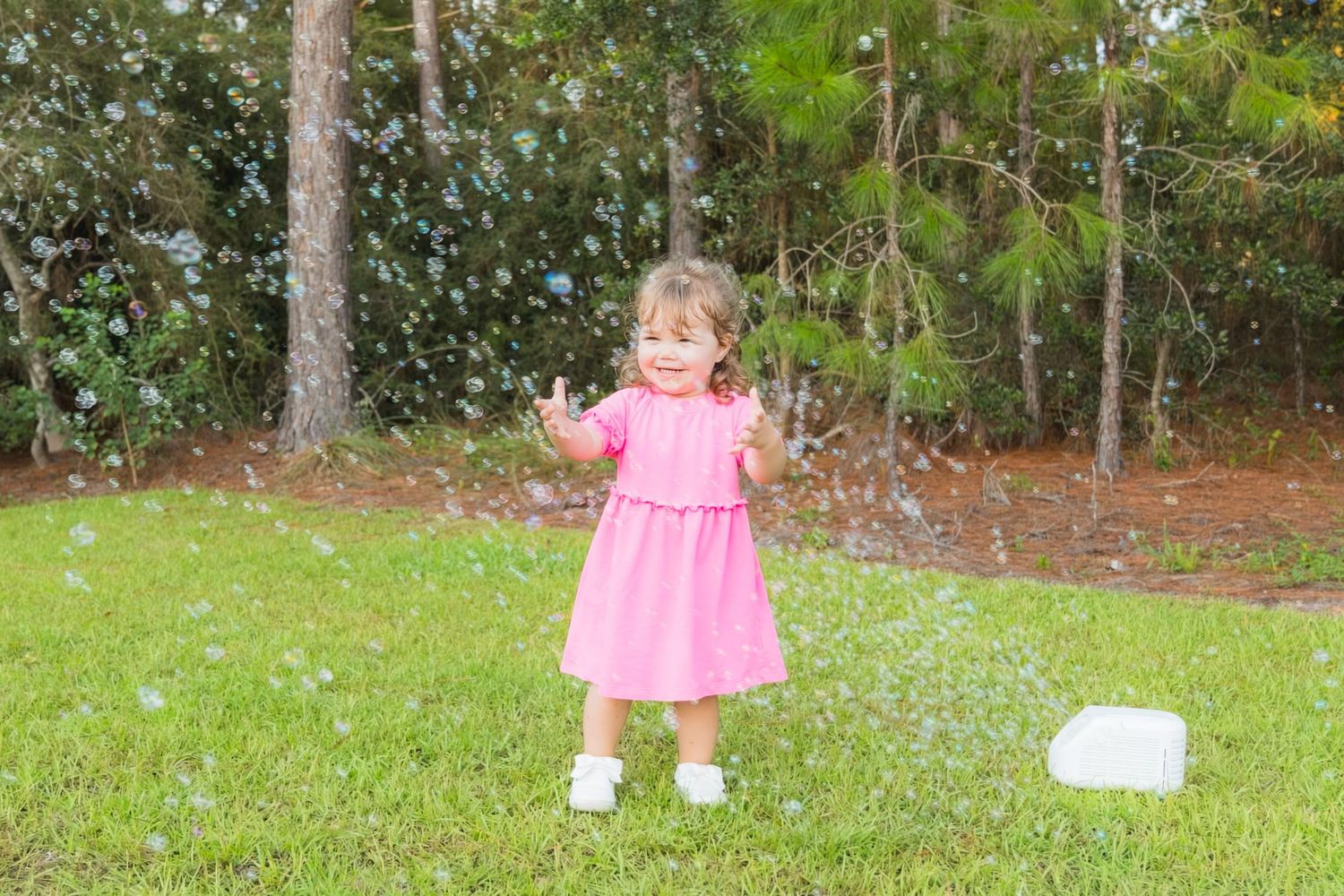 Toddler girl in pink dress playing with bubbles on green grass in a sunny backyard surrounded by trees.