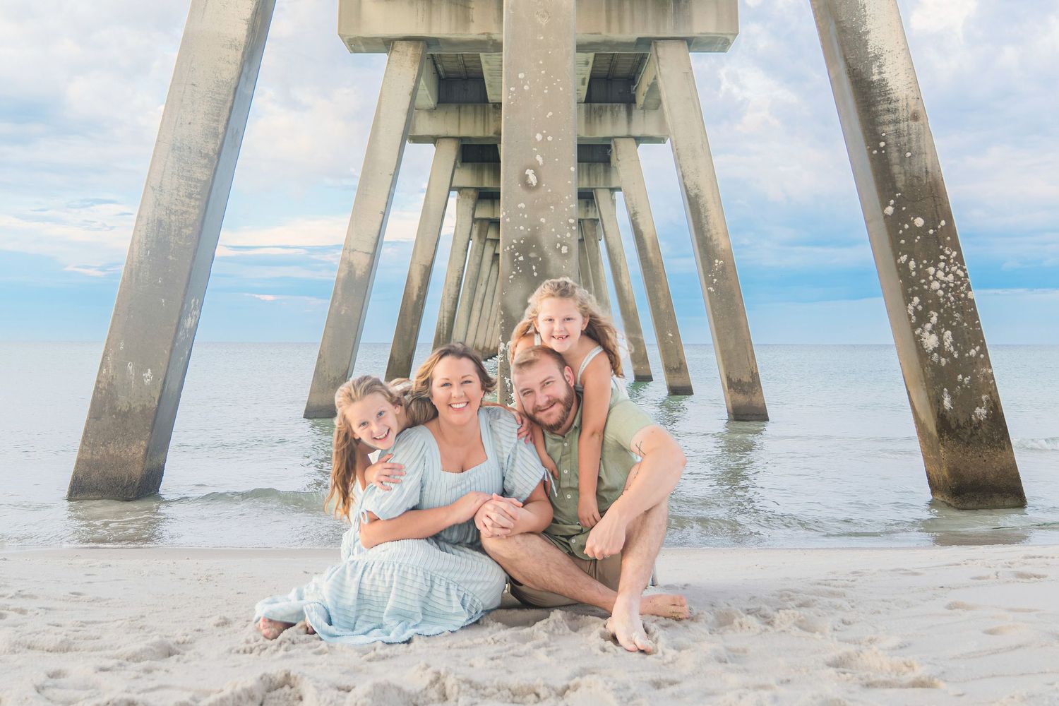 Family of four poses under a beach pier on sandy shore with calm blue ocean waters in the background.