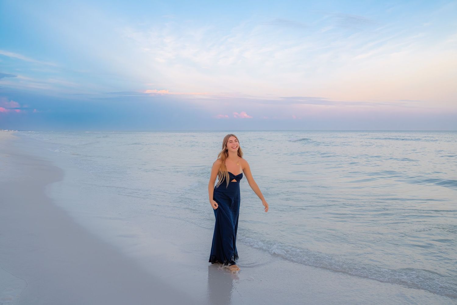 Woman in navy dress walking on white sand beach at sunset with pastel sky and calm ocean waves.