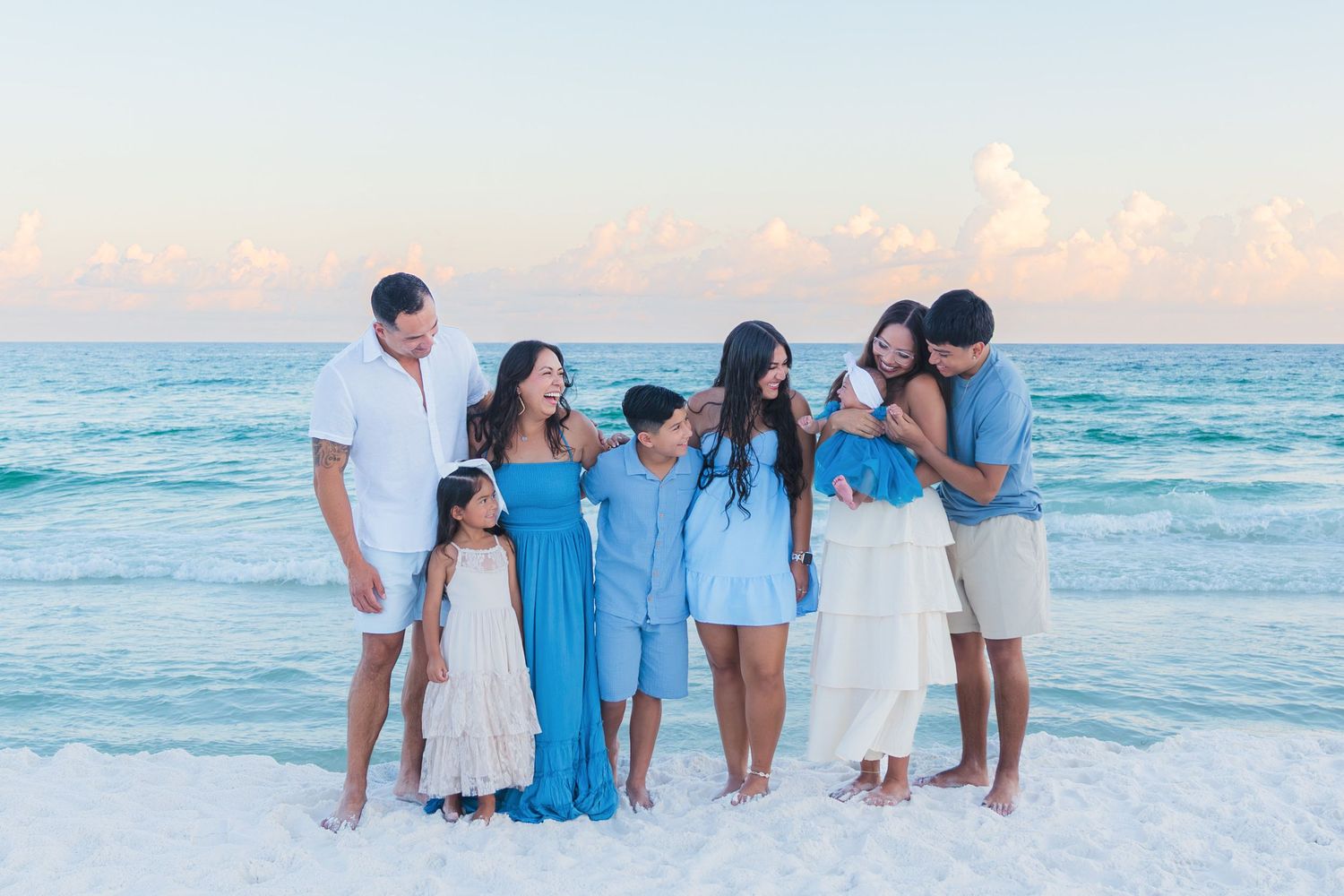 Family in blue and white outfits posing together on a white sand beach with turquoise water at sunset.