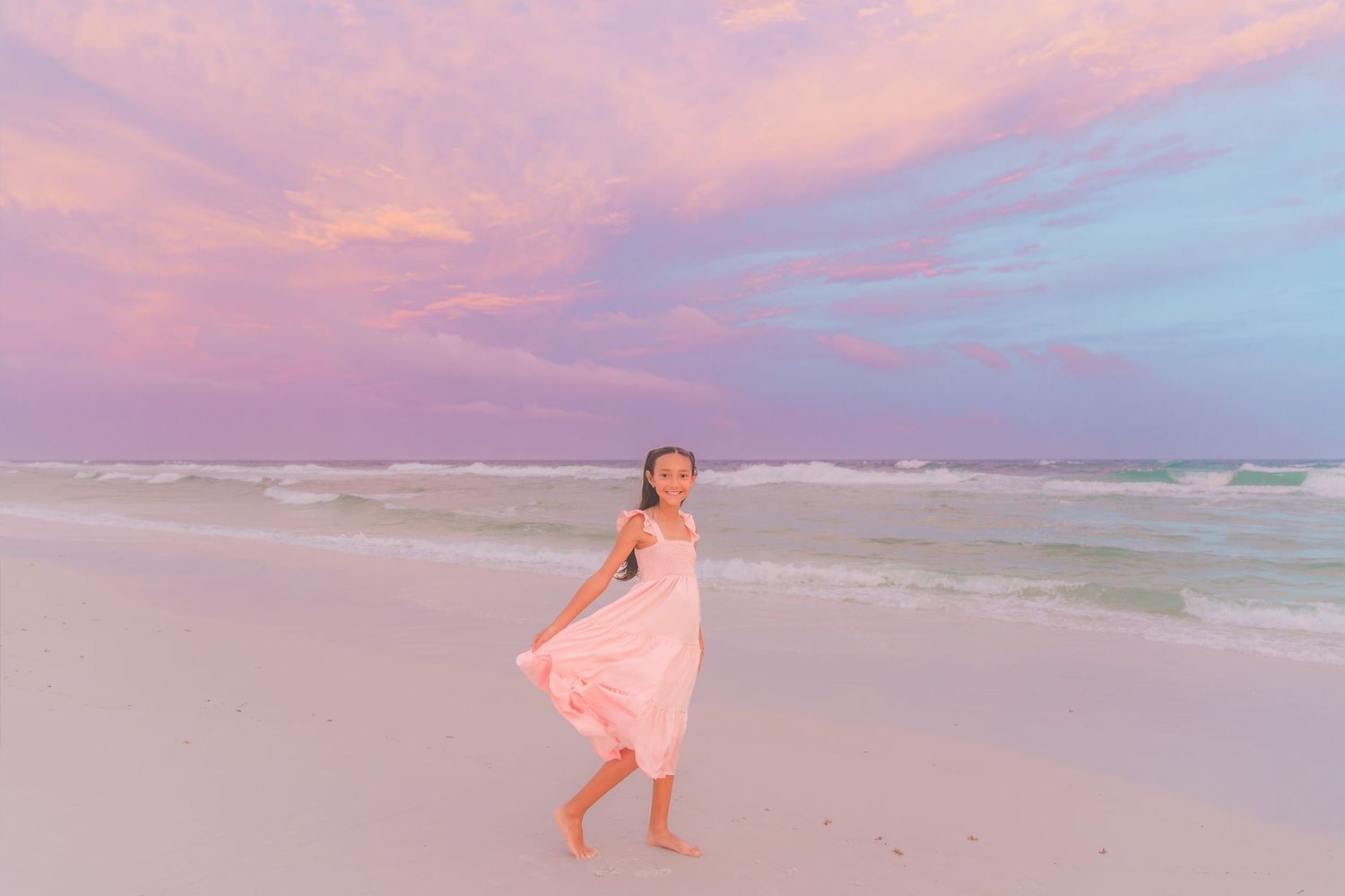 Young girl in pink dress twirling on a white sand beach at sunset with colorful pink and blue sky.