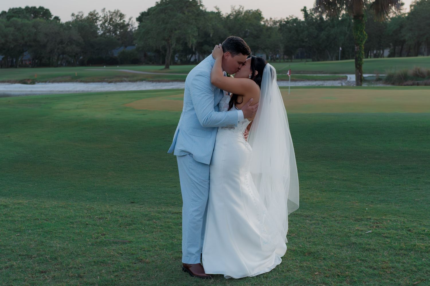 Bride and groom share first kiss on lush green golf course at golden hour outdoor wedding celebration.