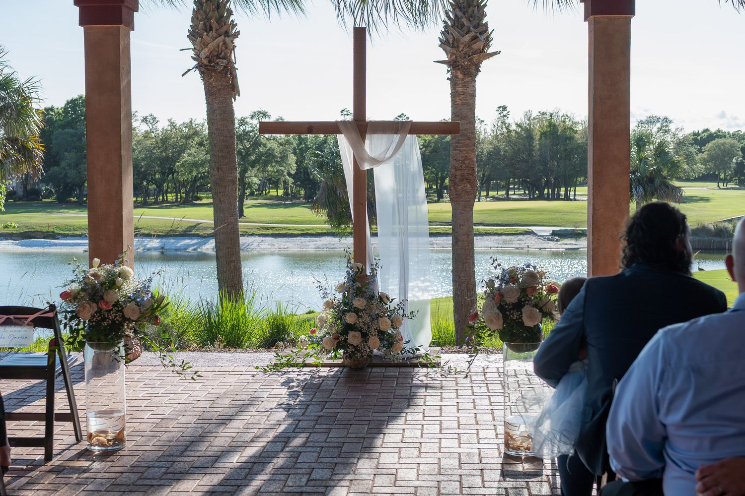 Outdoor wedding ceremony under wooden arch with floral arrangements, overlooking a serene lake and palm trees.