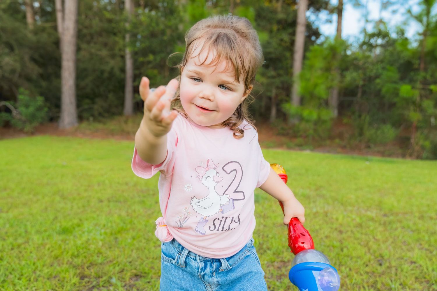 Toddler girl in pink unicorn shirt and jeans making a hand gesture while playing outdoors on green grass.