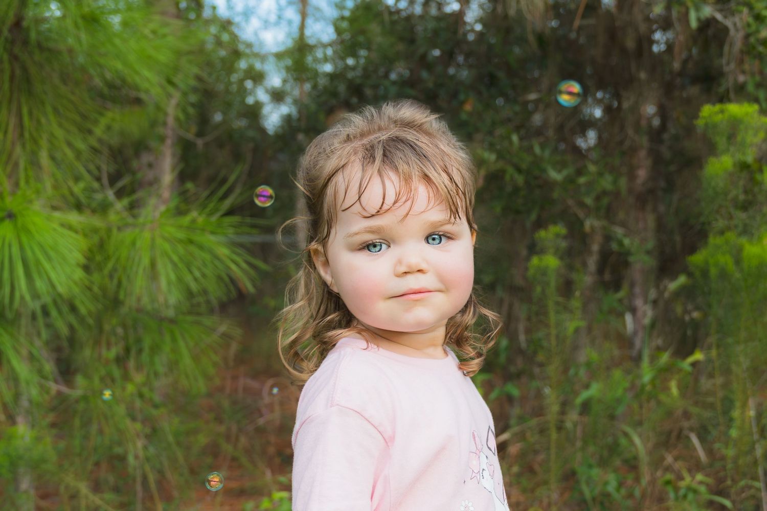 A young toddler girl with blue eyes and blonde hair wearing a pink shirt, standing outdoors near lush green trees.