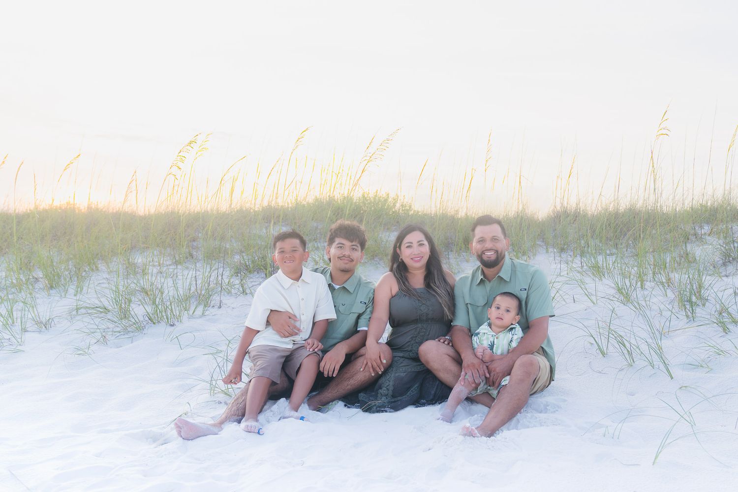 Family of five sitting together on white sand dunes with sea oats and soft sunset light.