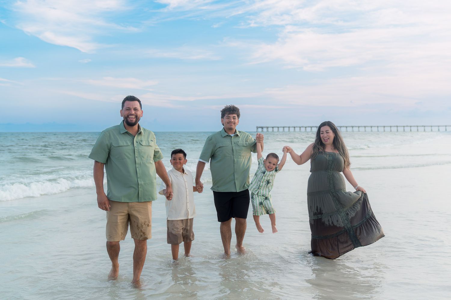 Family of five posing on a beach at sunset, wearing coordinating green outfits, with waves and a pier in the background.