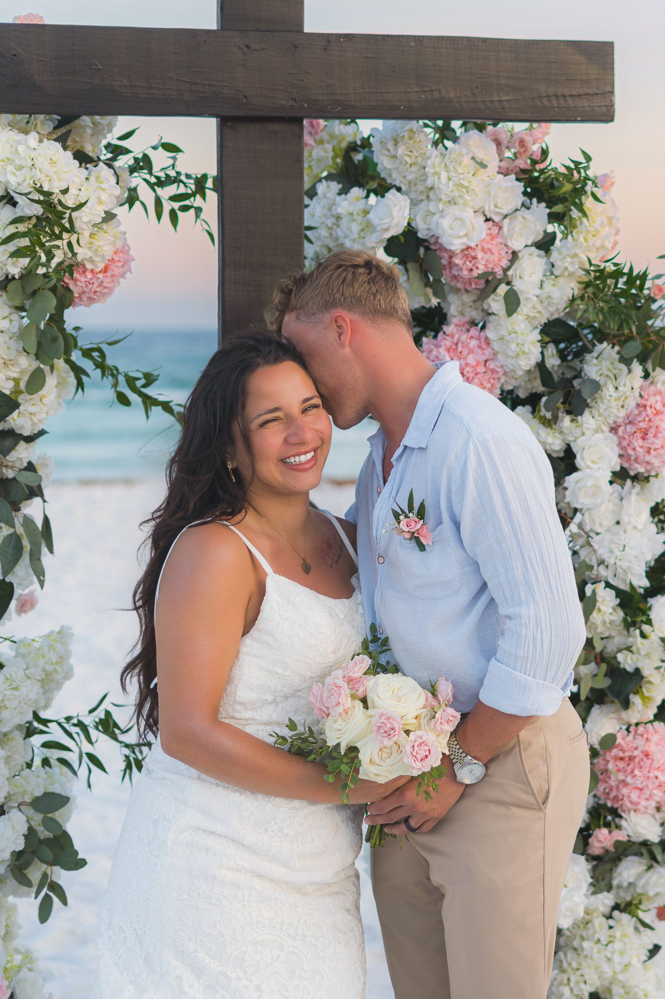 Bride smiles as groom kisses her forehead under a wooden cross adorned with white and pink hydrangeas on the beach.