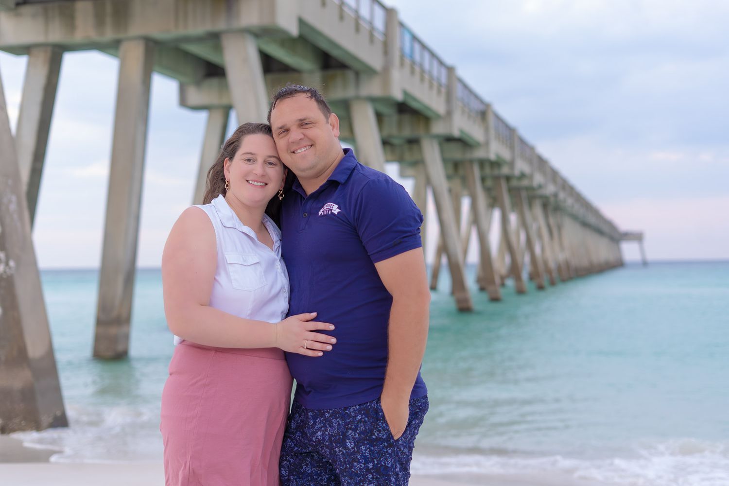 Couple embracing on a sandy beach near a wooden pier with turquoise ocean water in the background.