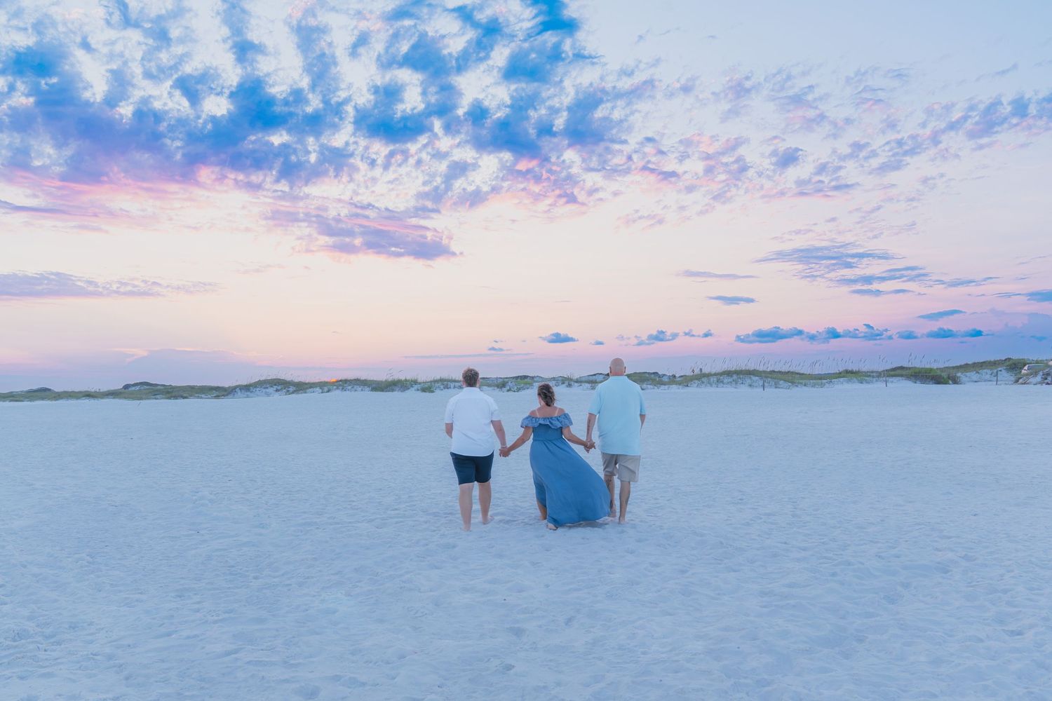 Family of three walking hand-in-hand on a white sandy beach at sunset with pastel pink and blue sky.