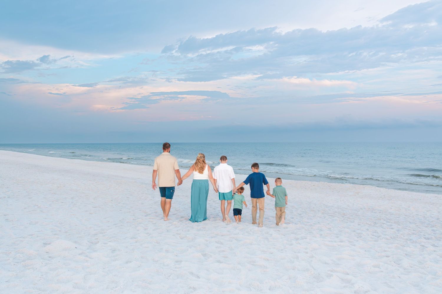 Family of five walking hand-in-hand on a white sandy beach at sunset with calm turquoise ocean waves.