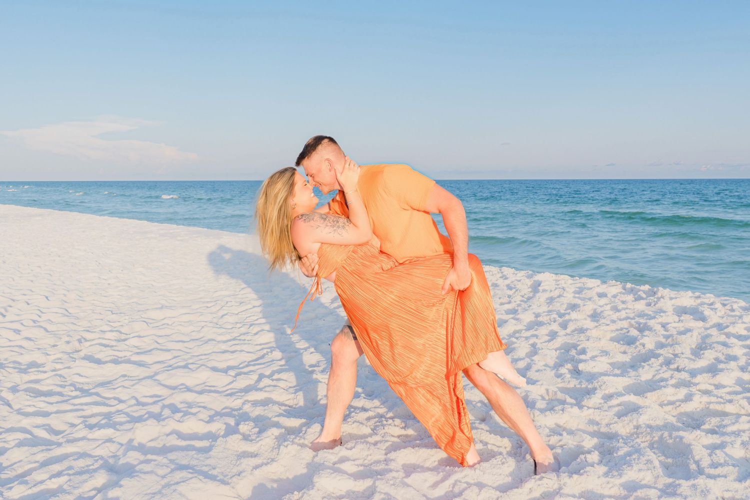 Couple in matching orange outfits sharing a romantic dip kiss on a white sand beach with turquoise ocean backdrop.