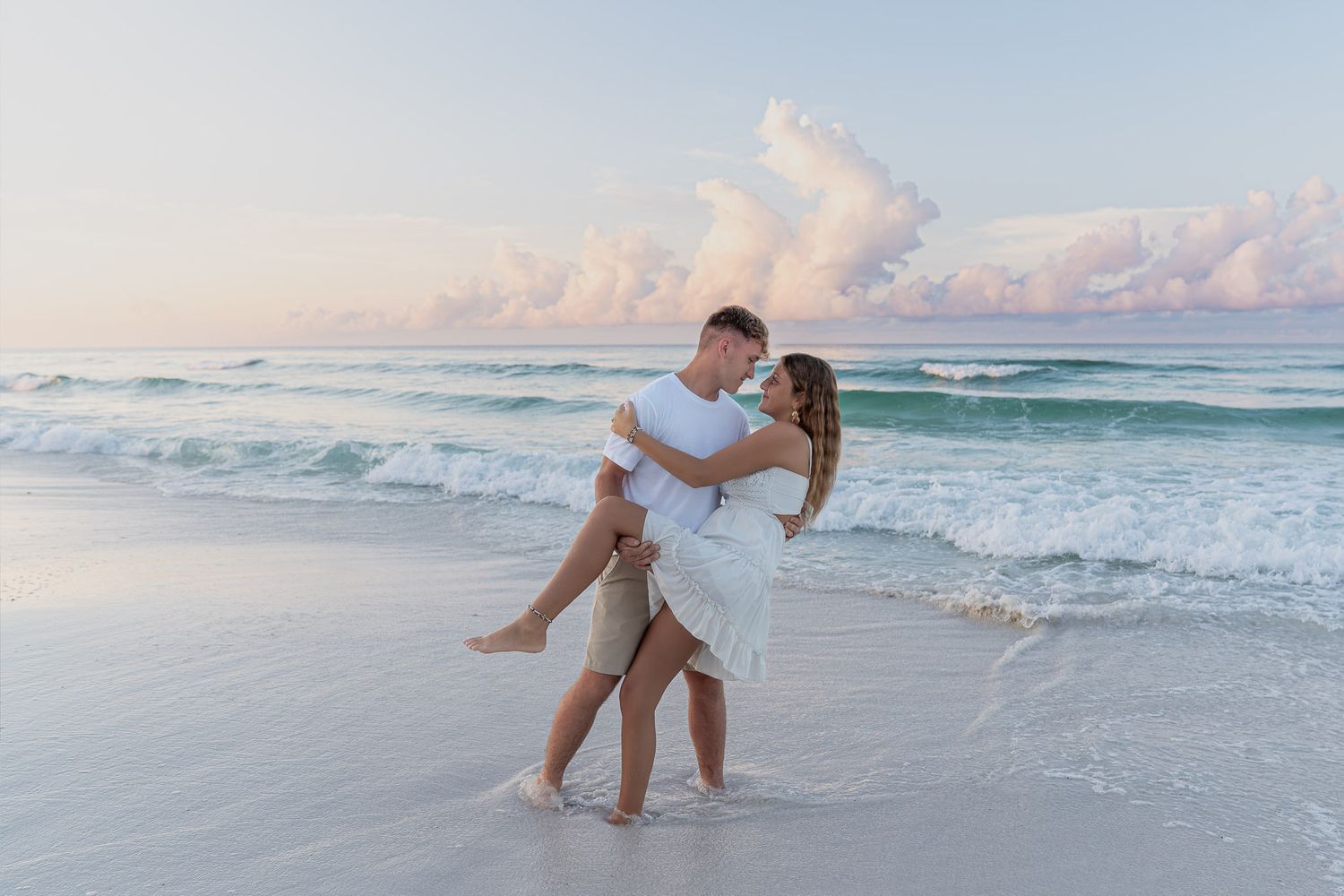 Couple embracing on a sandy beach at sunset, man lifting woman in white dress with turquoise ocean waves behind them.