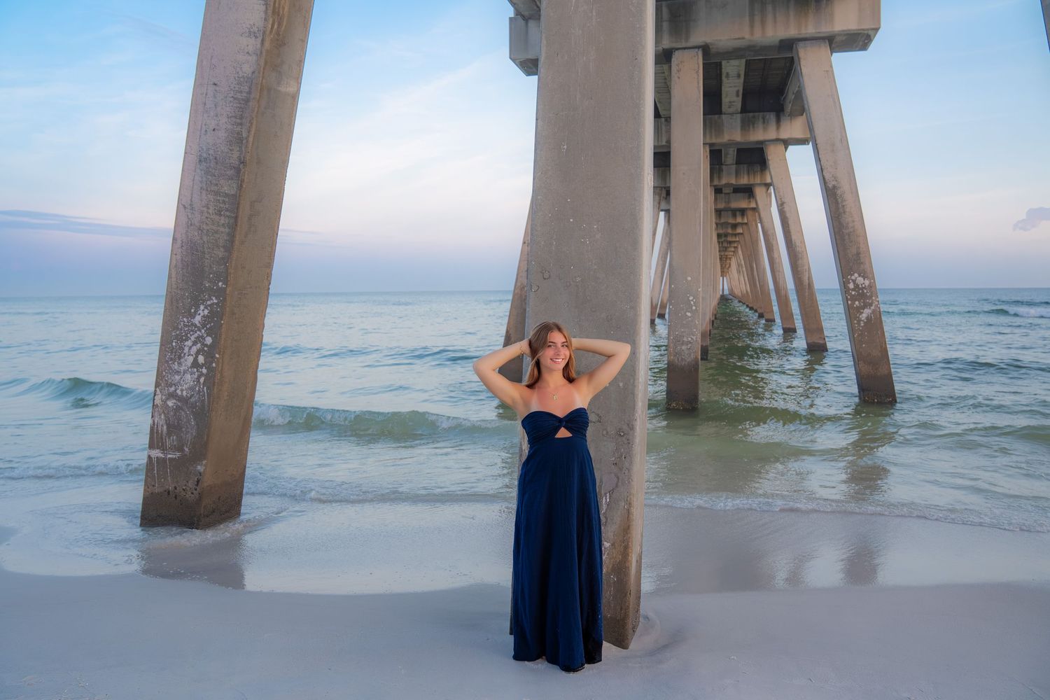Woman in navy blue gown poses under a beach pier at sunset on white sand with calm ocean waves.