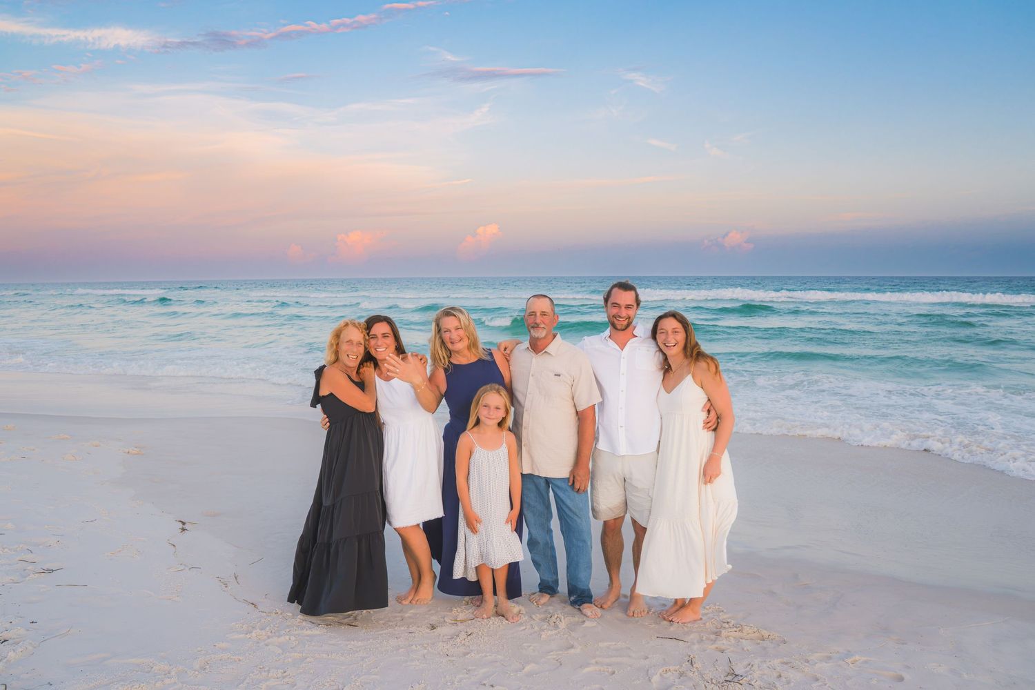 Family of seven posing together on a white sand beach at sunset with turquoise ocean waves behind them.