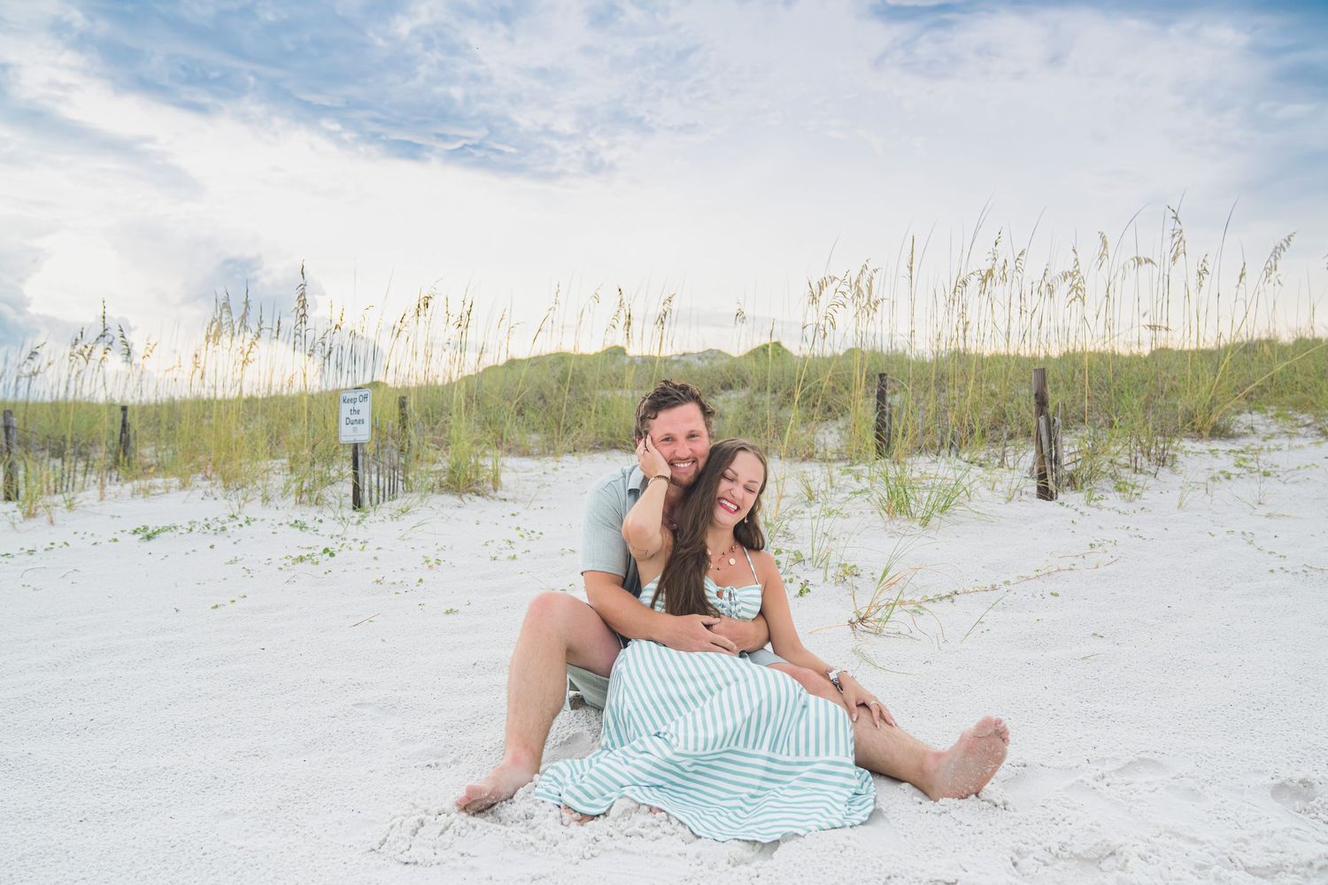 Happy couple sitting together on white sandy beach with sea oats and dunes in background.