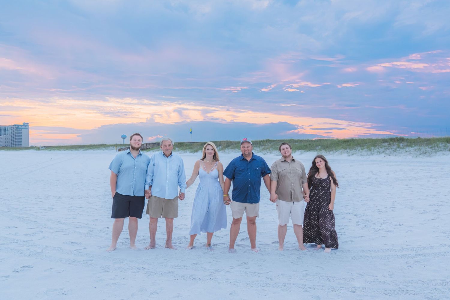 Group of six adults posing on a sandy beach at sunset with colorful pink and blue sky behind them.