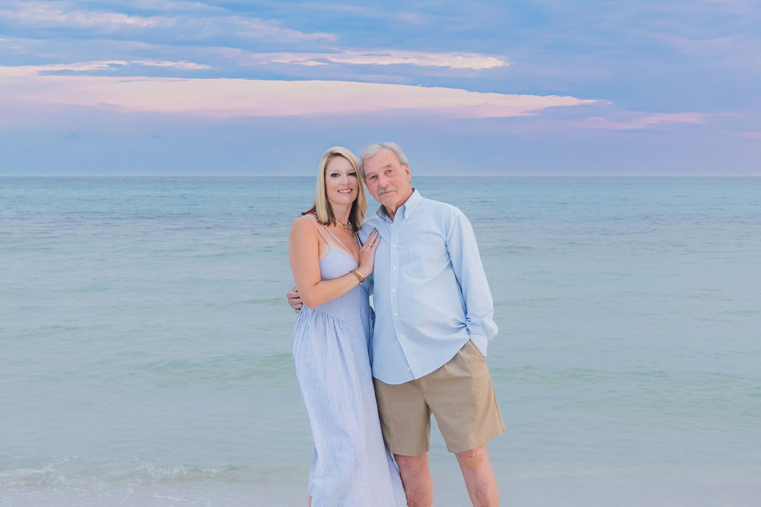 Couple posing together on a sandy beach at sunset with turquoise water and pastel sky behind them.