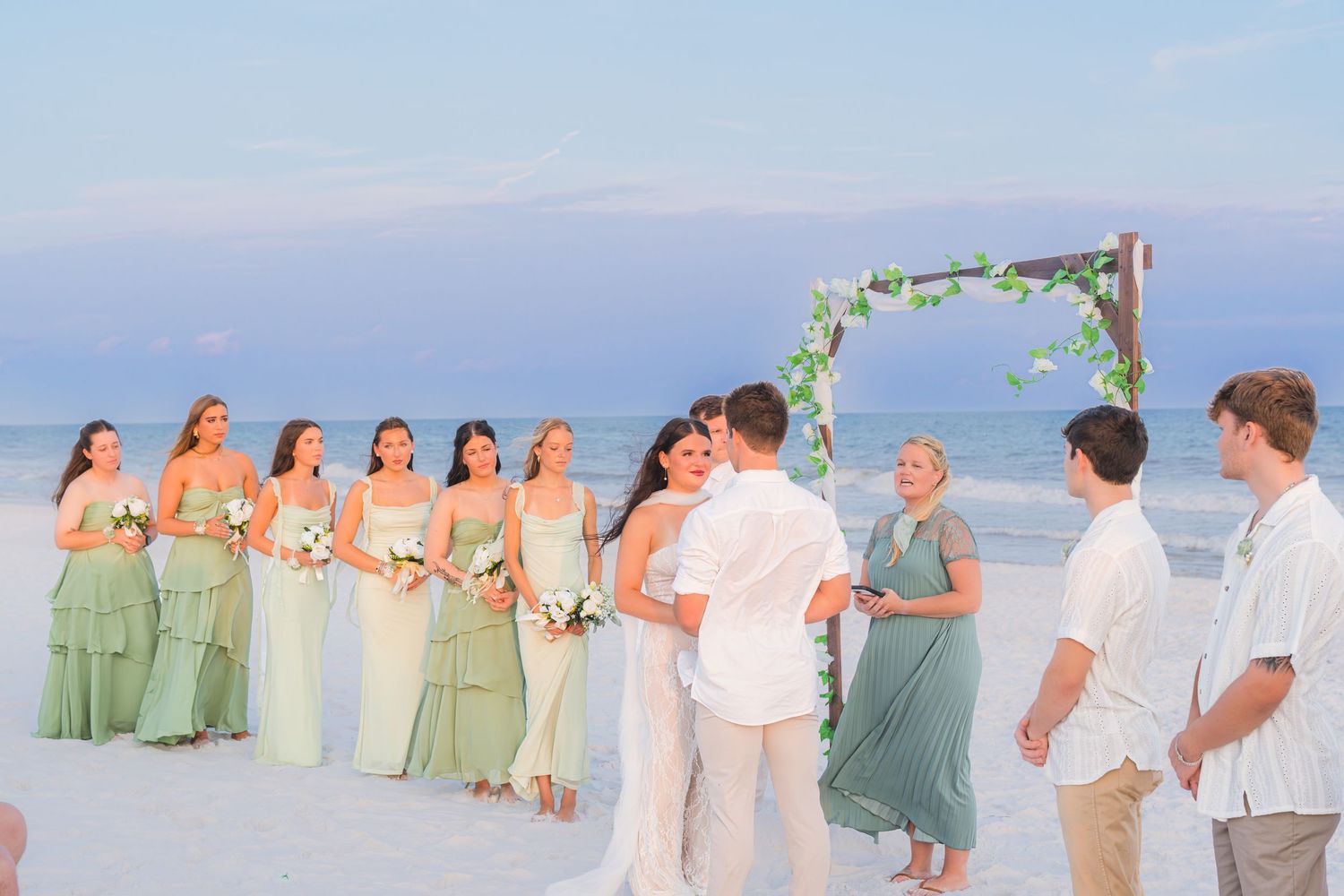 Beach wedding ceremony with bridesmaids in green dresses, couple kissing at floral arch, ocean backdrop at dusk.