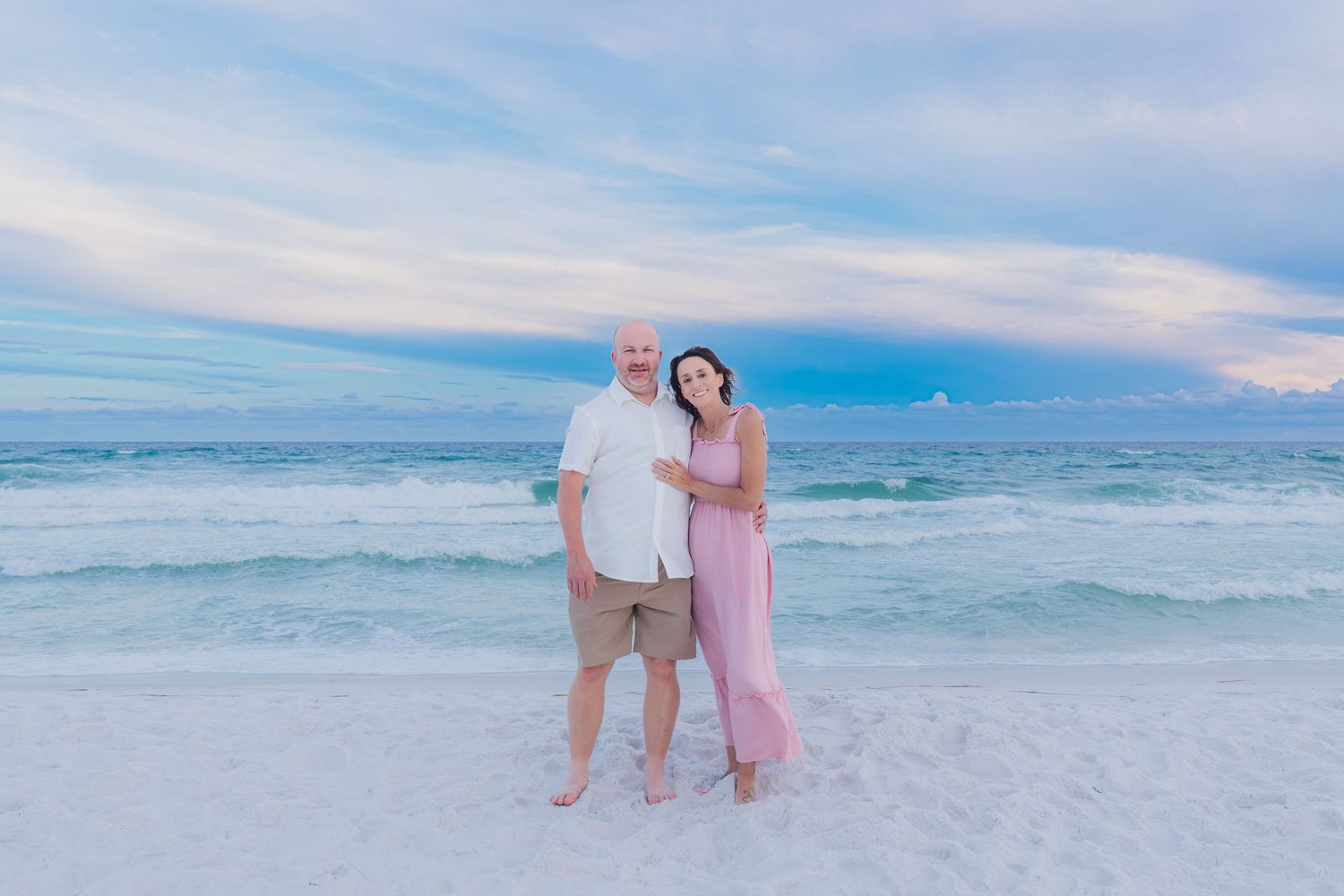 Couple posing together on a white sand beach at sunset with turquoise ocean waves behind them.