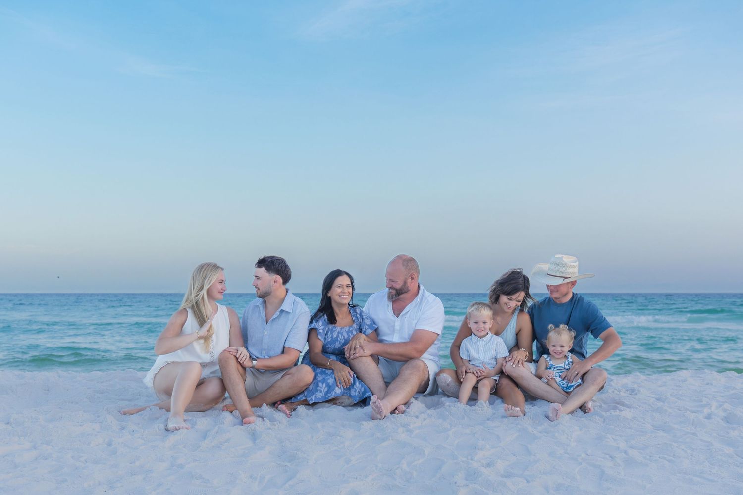 Family of seven poses on white sand beach at sunset with turquoise ocean waves in the background.