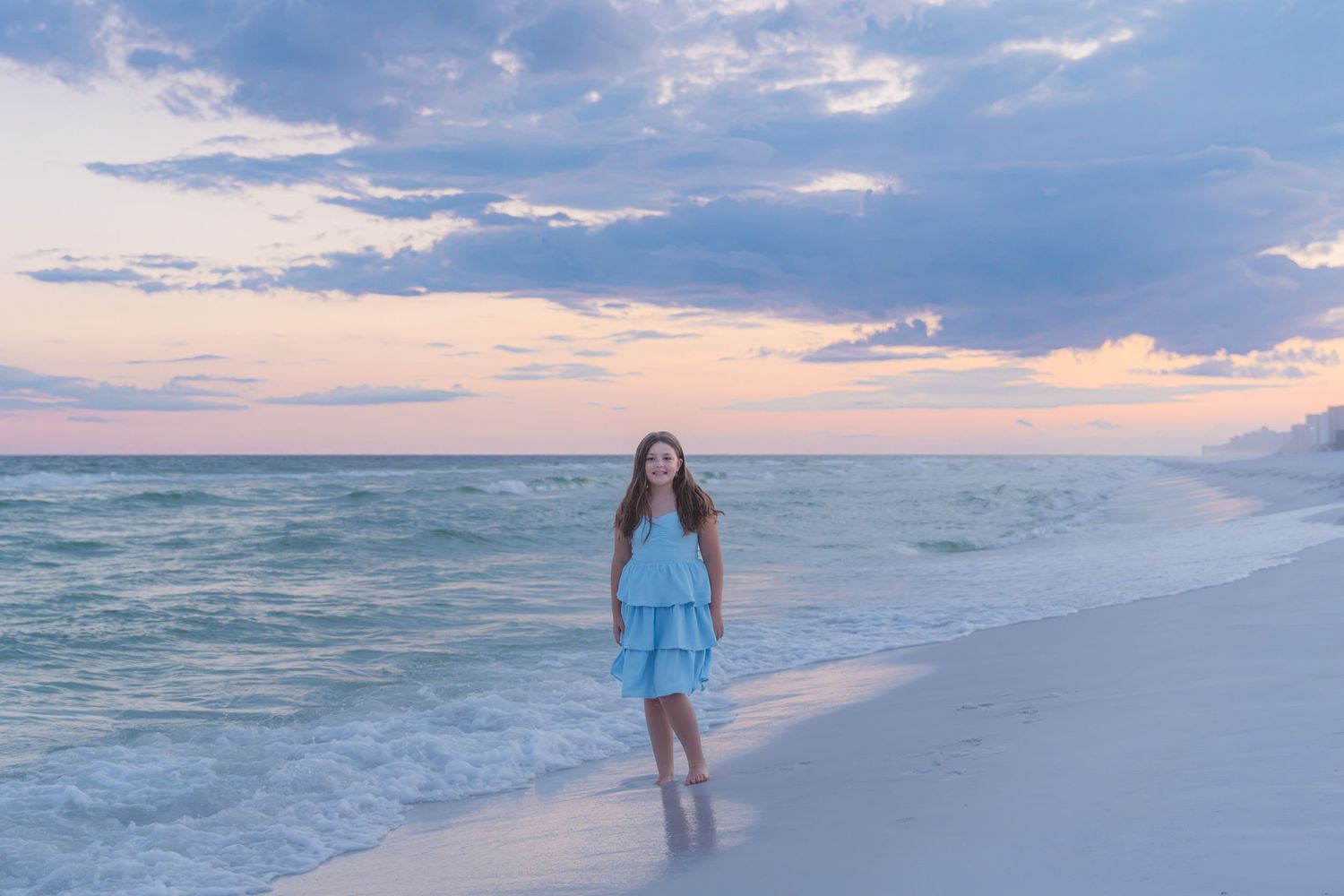 A young girl in a light blue dress walks along a sandy beach at sunset with soft waves and pastel skies.