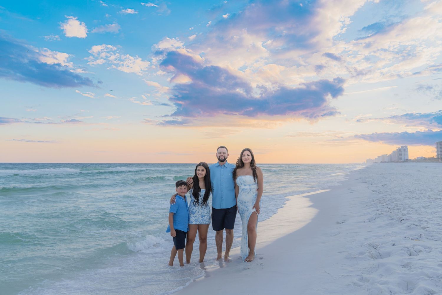 Family of four posing on a white sand beach at sunset with colorful pastel sky and turquoise ocean waves.