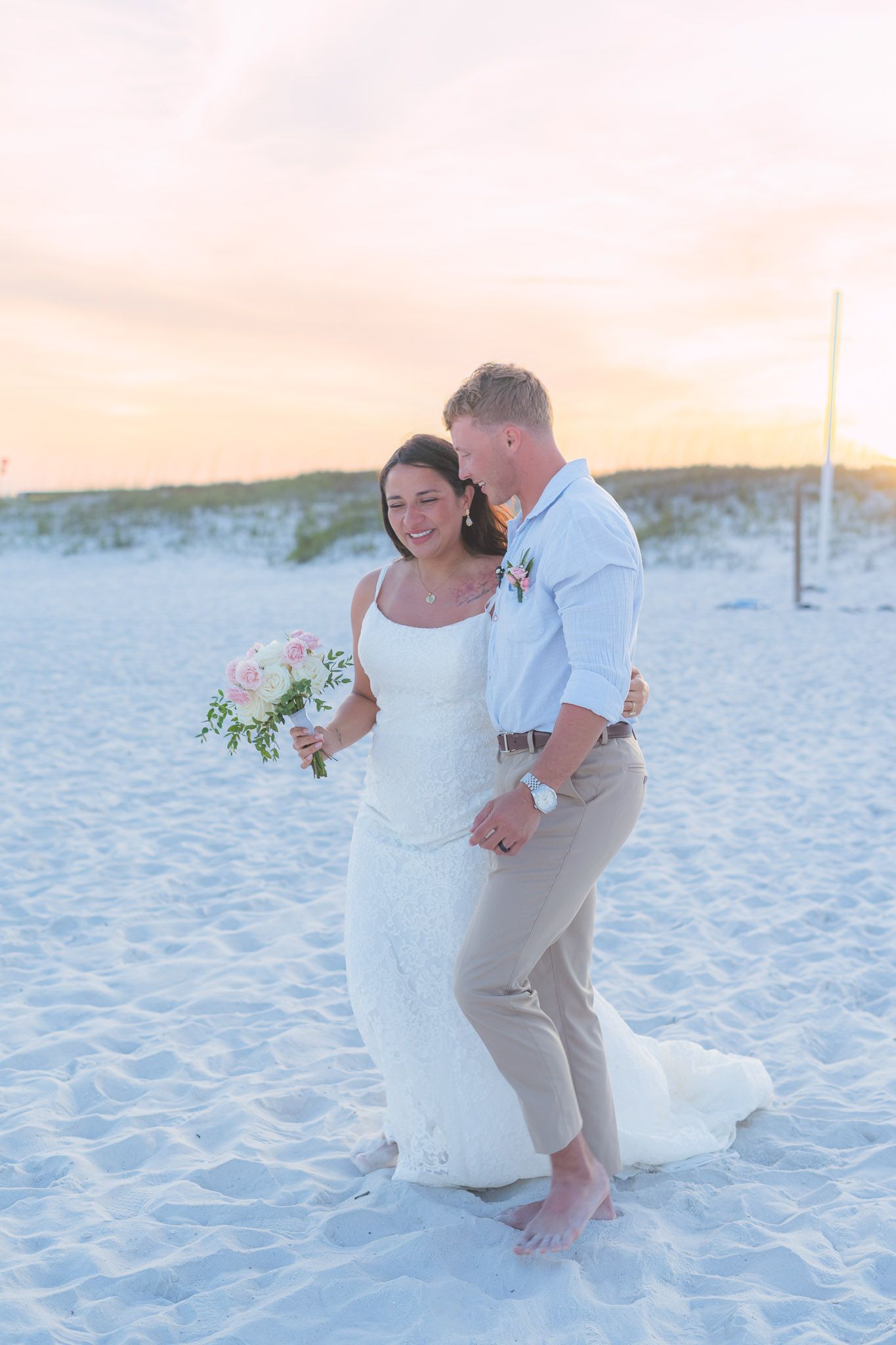 Happy newlyweds walk barefoot on white sand beach at golden sunset together.