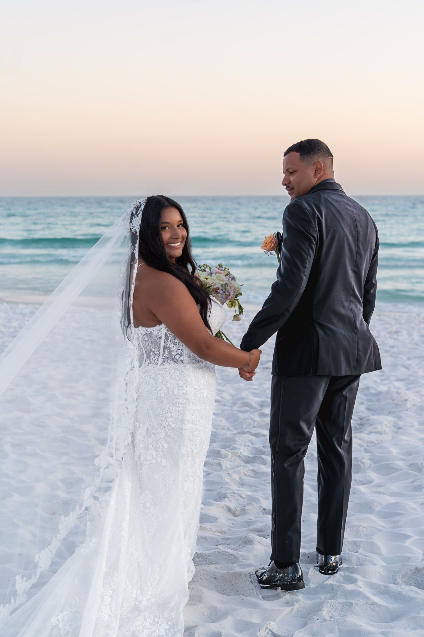 A bride and groom hold hands on a sandy beach at sunset, she smiles in a white gown with veil, he wears a dark suit.
